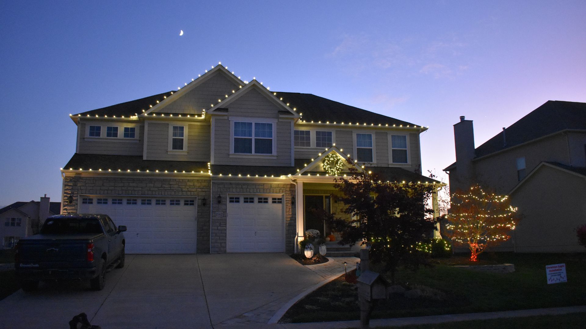 Suburban home at dusk with warm white holiday lights on the roofline and a tree wrapped in red and white lights in the front yard.